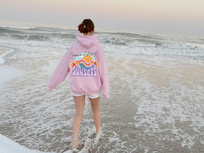 May include: A pink hoodie with the text "Forever Chasing Sunsets" and a graphic of a sunset with flowers on the back. The person wearing the hoodie is walking on a sandy beach with waves.