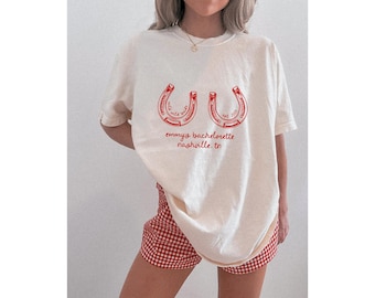 May include: Cream-colored t-shirt with a red horseshoe design and text that reads "emmy's bachelorette nashville, tn". The shirt is paired with red and white checkered shorts. The design includes the words "wild bride" and "est. 2023".