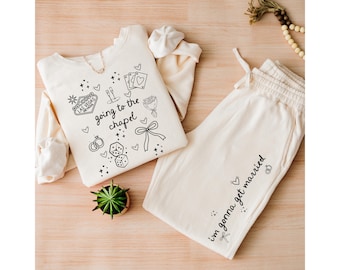 May include: A cream-coloured long-sleeve sweatshirt and matching drawstring sweatpants set. The sweatshirt features black line art of Las Vegas, playing cards, dice, rings, a bouquet, and the text "going to the chapel". The pants feature the text "i'm gonna get married".