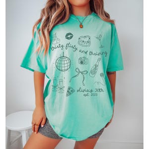 Może przedstawiać: Turkusowy t-shirt z nadrukiem przedstawiającym kulę dyskotekową, butelkę szampana, ciasto, wiśnie, aparat fotograficzny i tekst "thirty, flirty, and thriving. olivia's 30th est. 2023"