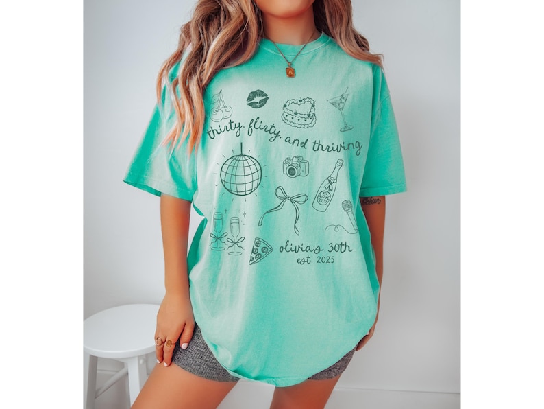 Może przedstawiać: Turkusowy t-shirt z nadrukiem przedstawiającym kulę dyskotekową, butelkę szampana, ciasto, wiśnie, aparat fotograficzny i tekst "thirty, flirty, and thriving. olivia's 30th est. 2023"