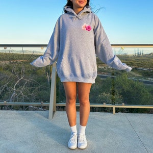 May include: A heather gray hoodie with a drawstring hood and a small floral design on the chest. The hoodie is worn with white sneakers and socks. The background features a scenic view with a clear blue sky.