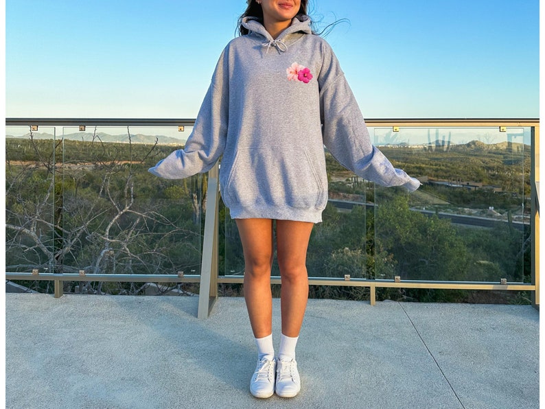 May include: A heather gray hoodie with a drawstring hood and a small floral design on the chest. The hoodie is worn with white sneakers and socks. The background features a scenic view with a clear blue sky.