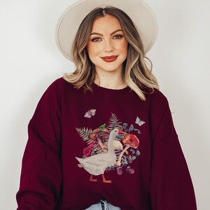 Puede incluir: Una sudadera color burdeos con un gr&aacute;fico de un ganso blanco y un hongo. El ganso est&aacute; rodeado de hojas, bayas y mariposas.