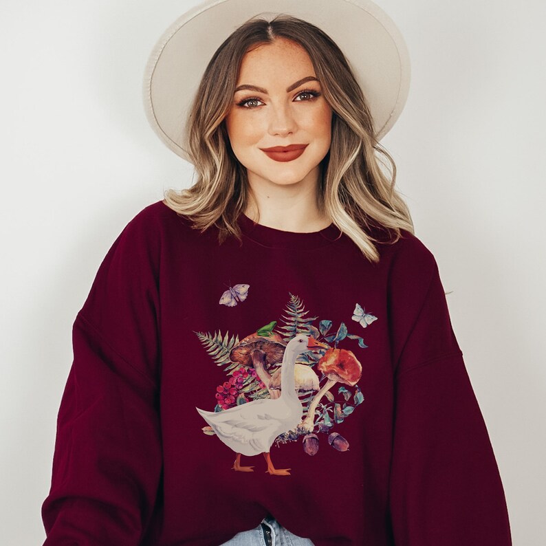 Puede incluir: Una sudadera color burdeos con un gr&aacute;fico de un ganso blanco y un hongo. El ganso est&aacute; rodeado de hojas, bayas y mariposas.