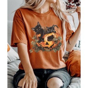 Peut inclure: Un t-shirt orange br&ucirc;l&eacute; avec un motif de trois chats noirs &agrave; l'int&eacute;rieur d'une citrouille.