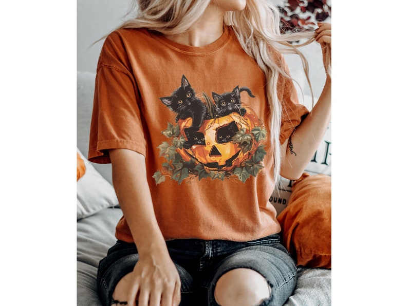 Peut inclure: Un t-shirt orange br&ucirc;l&eacute; avec un motif de trois chats noirs &agrave; l'int&eacute;rieur d'une citrouille.