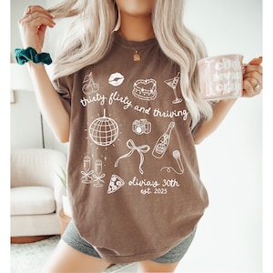 Może przedstawiać: Brązowy T-shirt z białym tekstem, kt&oacute;ry brzmi "thirty flirty and thriving" i zawiera r&oacute;żne ilustracje, w tym kulę dyskotekową, butelkę szampana i kawałek pizzy. Tekst zawiera r&oacute;wnież napis "Olivia's 30th est. 2023".