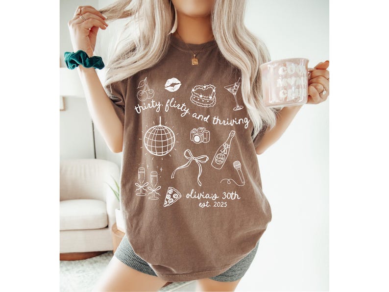 Może przedstawiać: Brązowy T-shirt z białym tekstem, kt&oacute;ry brzmi "thirty flirty and thriving" i zawiera r&oacute;żne ilustracje, w tym kulę dyskotekową, butelkę szampana i kawałek pizzy. Tekst zawiera r&oacute;wnież napis "Olivia's 30th est. 2023".