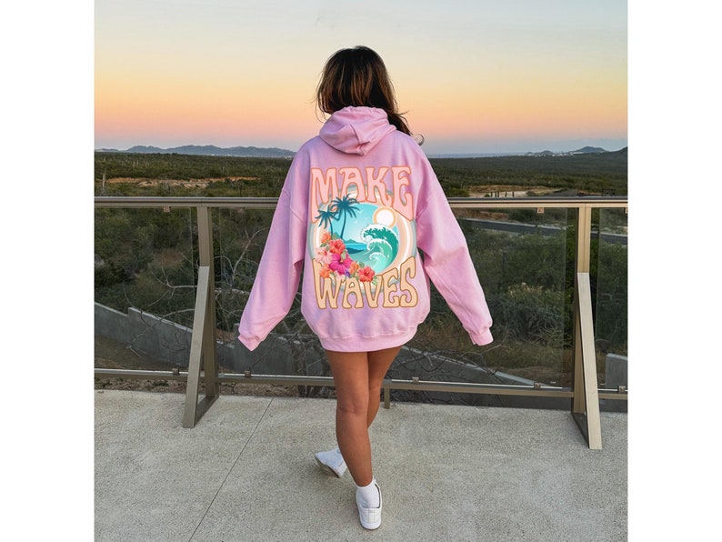 May include: A light pink hoodie with the words "MAKE WAVES" in a retro font. The back of the hoodie features a graphic of a tropical scene with palm trees, waves, and flowers. The hoodie is worn outdoors.