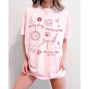 Może przedstawiać: R&oacute;żowa koszulka z nadrukiem graficznym, kt&oacute;ry głosi "thirty flirty and thriving" z r&oacute;żnymi ilustracjami, w tym dyskoteką, butelką szampana, kawałkiem pizzy i wiśniami. Tekst "Olivia's 30th est. 2025" jest r&oacute;wnież zawarty w projekcie.