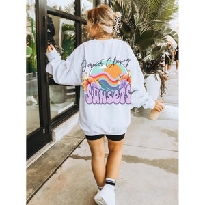 May include: White sweatshirt with a graphic print of a sunset with the text "Forever Chasing Sunsets" in purple and white.
