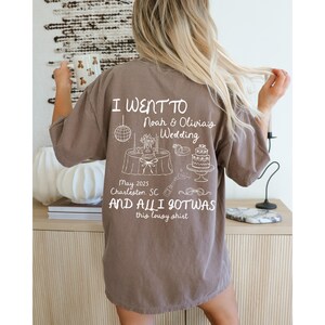 May include: A brown t-shirt with white text that reads "I went to Noah & Olivia's Wedding May 2025 Charleston, SC and all I got was this lousy shirt". The shirt has a small illustration of a wedding cake, a table with a disco ball, and other wedding-related items.
