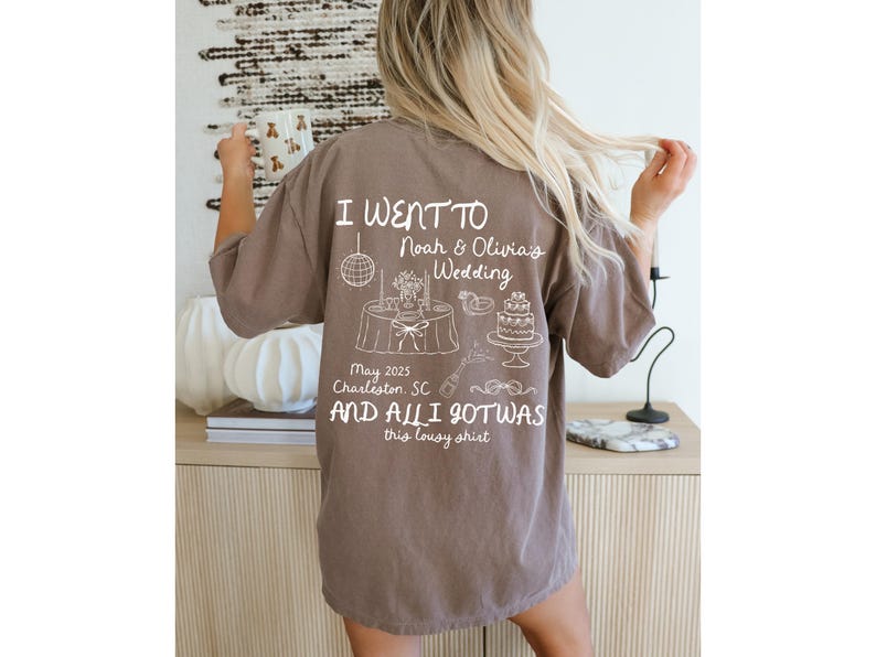 May include: A brown t-shirt with white text that reads "I went to Noah & Olivia's Wedding May 2025 Charleston, SC and all I got was this lousy shirt". The shirt has a small illustration of a wedding cake, a table with a disco ball, and other wedding-related items.