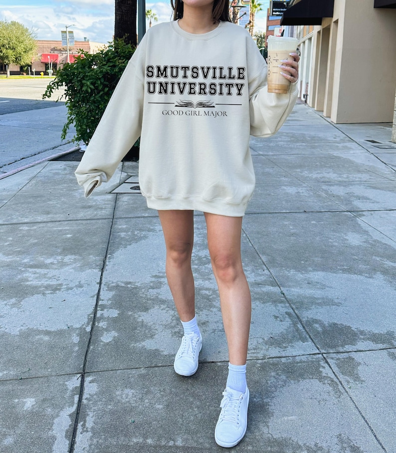 May include: A cream-colored sweatshirt with the text "Smutsville University" and "Good Girl Major" printed in black. The sweatshirt is worn by a person standing on a sidewalk.