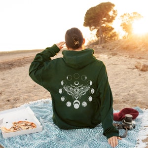 May include: Dark green hoodie with a white moth and moon phase design on the back. The person is sitting on a blanket on the beach with a pizza box and a camera.