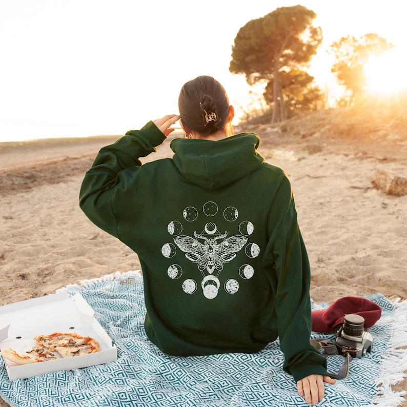 May include: Dark green hoodie with a white moth and moon phase design on the back. The person is sitting on a blanket on the beach with a pizza box and a camera.