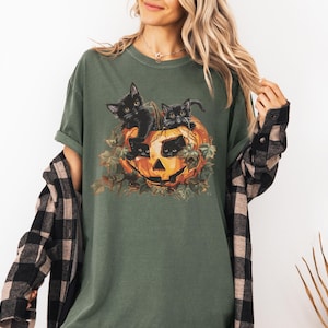 Peut inclure: Un t-shirt vert avec un motif de trois chatons noirs &agrave; l'int&eacute;rieur d'une citrouille. Les chatons regardent hors de la citrouille.