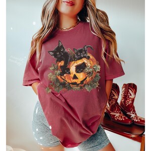 Peut inclure: Un t-shirt marron avec un motif de trois chatons noirs &agrave; l'int&eacute;rieur d'une citrouille d'Halloween. Les chatons regardent hors de la citrouille.