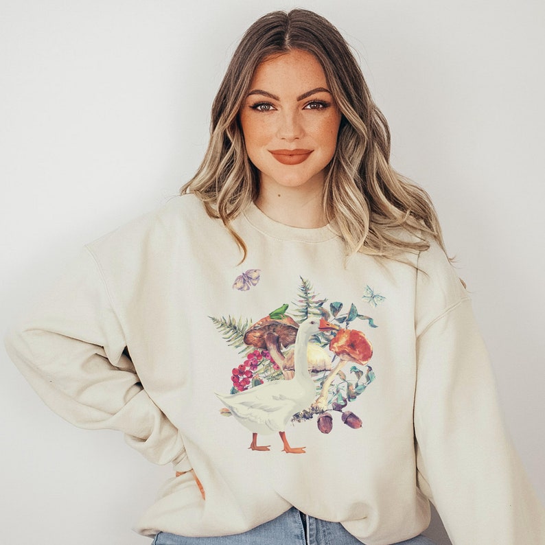 Puede incluir: Una sudadera de color crema con un estampado gr&aacute;fico de un ganso blanco rodeado de setas, hojas y mariposas de colores.