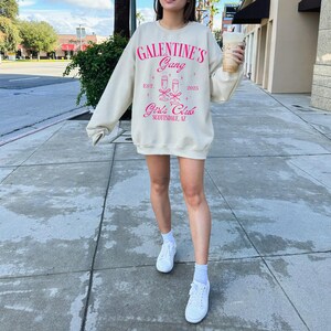 May include: A light beige sweatshirt with the text "Galentine's Gang" in pink lettering. The sweatshirt also features a graphic of two champagne glasses with pink bows and the text "Girls Club Scottsdale, AZ" in pink lettering.