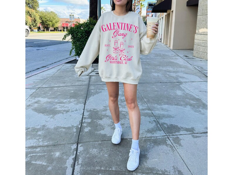 May include: A light beige sweatshirt with the text "Galentine's Gang" in pink lettering. The sweatshirt also features a graphic of two champagne glasses with pink bows and the text "Girls Club Scottsdale, AZ" in pink lettering.