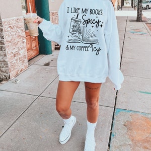 May include: White sweatshirt with black text that reads "I like my books spicy & my coffee icy". The text is accompanied by a graphic of a hand holding a cup of coffee and a book.