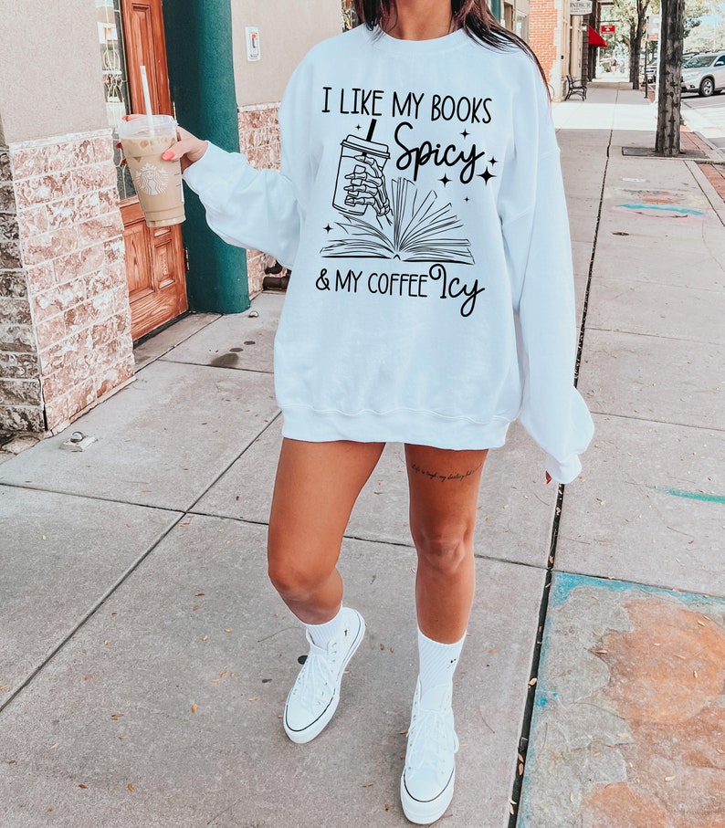 May include: White sweatshirt with black text that reads "I like my books spicy & my coffee icy". The text is accompanied by a graphic of a hand holding a cup of coffee and a book.