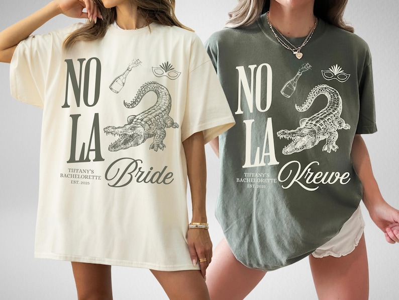 Two oversized t-shirts, one ivory and one moss, featuring a graphic design. The design includes the text "NO LA", an alligator, a champagne bottle, and a mask. The ivory shirt says "Bride", and the moss shirt says "Krewe".