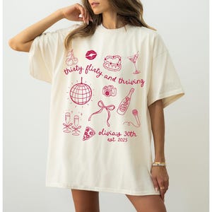 Może przedstawiać: Biały t-shirt z r&oacute;żowym nadrukiem graficznym, na kt&oacute;rym widnieje napis "thirty flirty and thriving" oraz ilustracje dyskotekowej kuli, kieliszk&oacute;w do szampana, kawałka pizzy, aparatu fotograficznego, szminki, wiśni, kieliszka do martini i mikrofonu. W projekcie znajduje się r&oacute;wnież tekst "Olivia's 30th est. 2023".