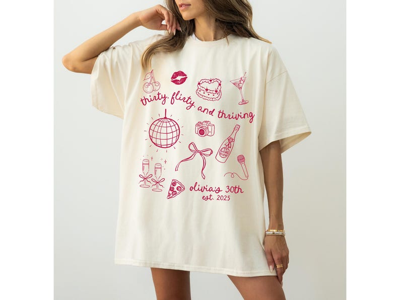 Może przedstawiać: Biały t-shirt z r&oacute;żowym nadrukiem graficznym, na kt&oacute;rym widnieje napis "thirty flirty and thriving" oraz ilustracje dyskotekowej kuli, kieliszk&oacute;w do szampana, kawałka pizzy, aparatu fotograficznego, szminki, wiśni, kieliszka do martini i mikrofonu. W projekcie znajduje się r&oacute;wnież tekst "Olivia's 30th est. 2023".