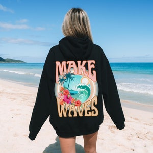 May include: Black hoodie with a colorful graphic on the back. The graphic features the text "MAKE WAVES" above a tropical scene with palm trees, a wave, and flowers. The hoodie is worn on a beach.