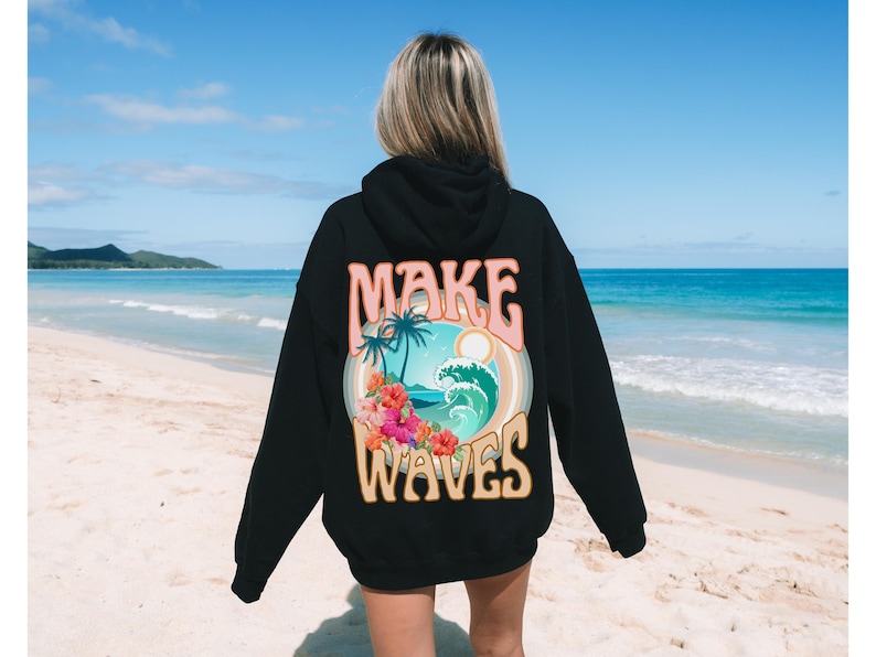 May include: Black hoodie with a colorful graphic on the back. The graphic features the text "MAKE WAVES" above a tropical scene with palm trees, a wave, and flowers. The hoodie is worn on a beach.