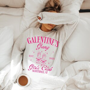 May include: A white sweatshirt with the text "Galentine's Gang Est. 2025 Girl's Club Scottsdale, AZ" printed in pink. The sweatshirt features a graphic of two champagne glasses with a pink ribbon.
