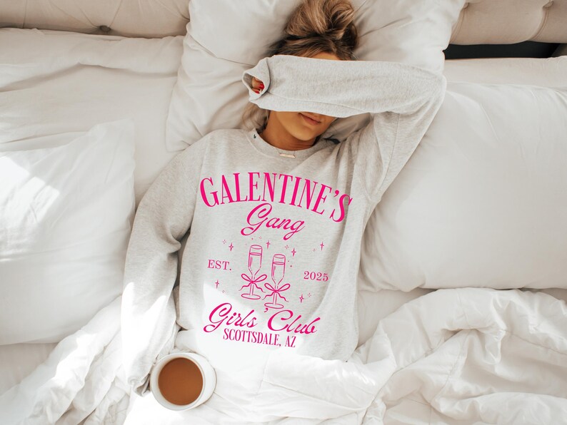 May include: A white sweatshirt with the text "Galentine's Gang Est. 2025 Girl's Club Scottsdale, AZ" printed in pink. The sweatshirt features a graphic of two champagne glasses with a pink ribbon.