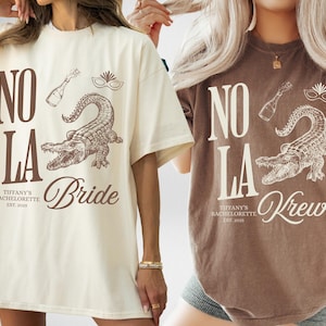 May include: Two oversized t-shirts, one cream and one brown, featuring a New Orleans-themed design. The design includes the text "NO LA", an alligator, champagne bottle, and mask. The cream shirt says "Bride", and the brown shirt says "Krewe".