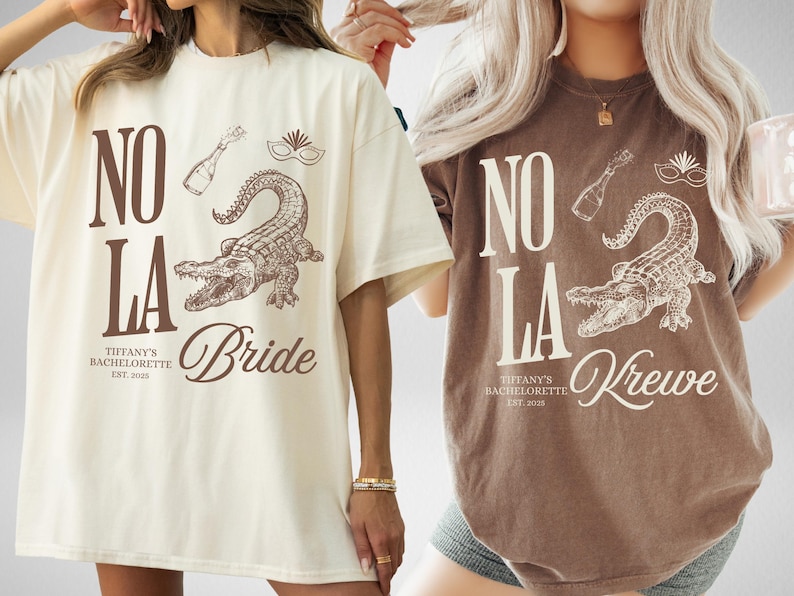 May include: Two oversized t-shirts, one cream and one brown, featuring a New Orleans-themed design. The design includes the text "NO LA", an alligator, champagne bottle, and mask. The cream shirt says "Bride", and the brown shirt says "Krewe".