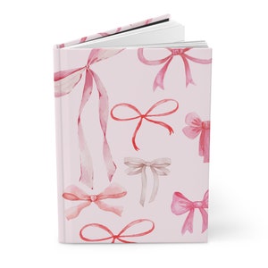 May include: A hardcover journal with a light pink cover adorned with watercolor illustrations of various pink and beige bows. The journal is open, revealing blank white pages.