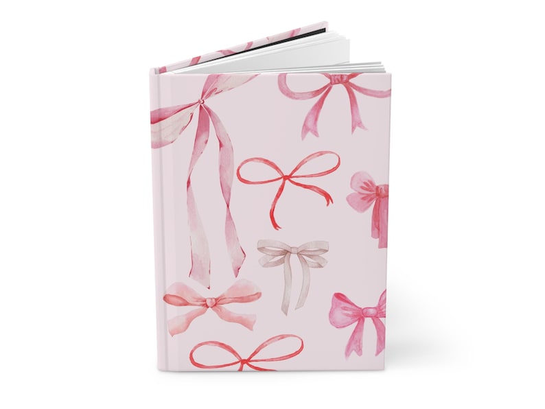 May include: A hardcover journal with a light pink cover adorned with watercolor illustrations of various pink and beige bows. The journal is open, revealing blank white pages.