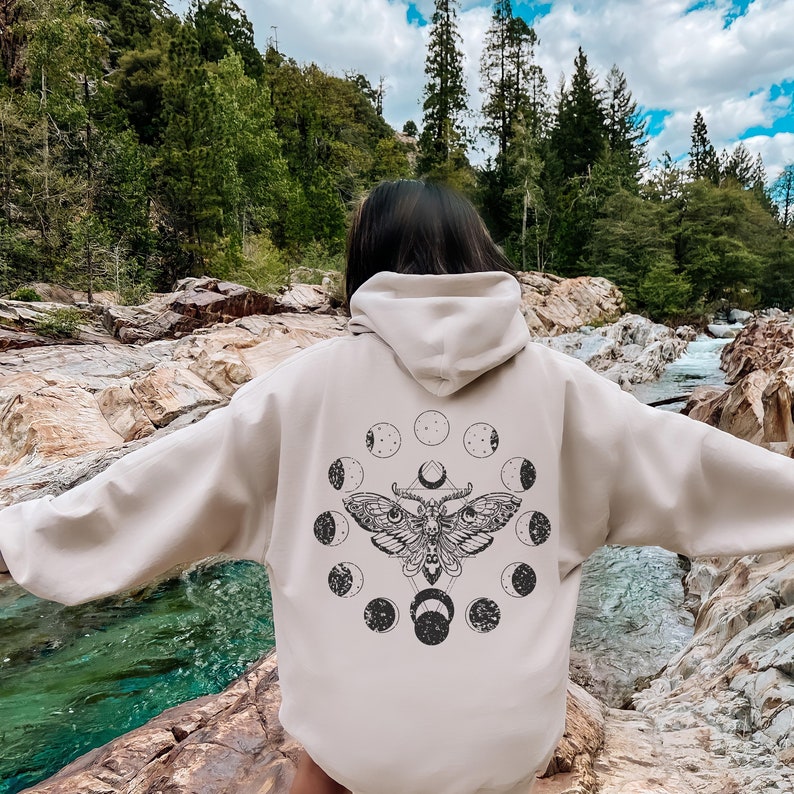 May include: Beige hoodie with a black moth and moon phase design on the back. The graphic features a detailed moth illustration surrounded by circular moon phases. The hoodie has a hood and long sleeves, suitable for casual wear.