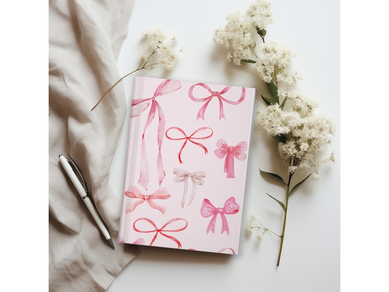 May include: A pink journal with a pattern of watercolor bows in shades of pink and red. A white pen rests on a beige cloth next to the journal. White flowers are arranged on a white surface.