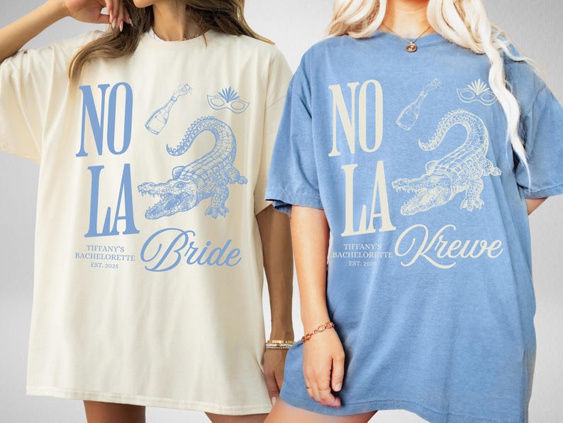 May include: Two oversized t-shirts, one ivory and one blue jean, featuring a light blue alligator graphic and the text "NO LA". The ivory shirt says "Bride", and the blue jean shirt says "Krewe". Both shirts have "Tiffany's Bachelorette" printed below.