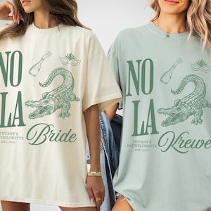 Two oversized t-shirts, one ivory and one bay, featuring a green graphic design. The design includes the text "NO LA", a champagne bottle, a mask, and an alligator. The ivory shirt says "Bride", and the bay shirt says "Krewe".