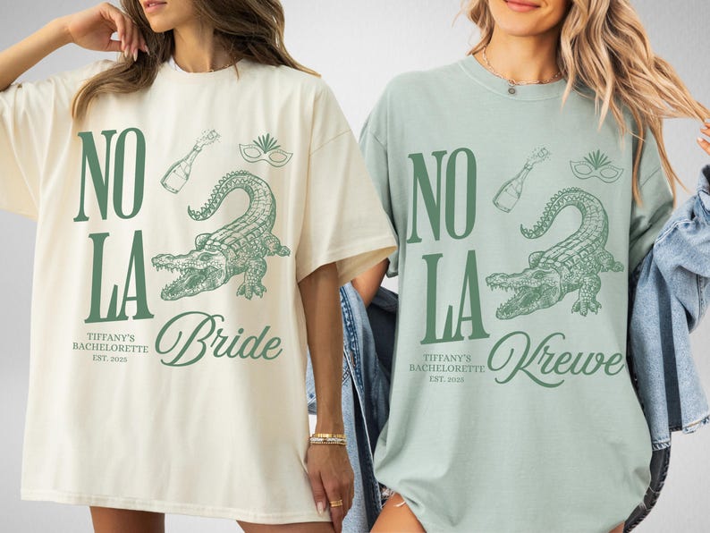 Two oversized t-shirts, one ivory and one bay, featuring a green graphic design. The design includes the text "NO LA", a champagne bottle, a mask, and an alligator. The ivory shirt says "Bride", and the bay shirt says "Krewe".