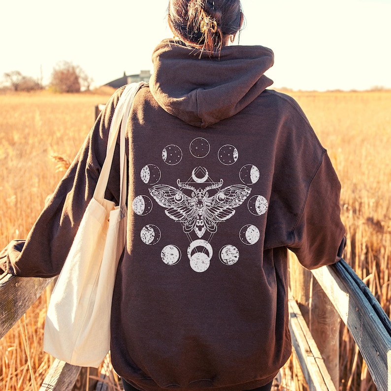 May include: A dark brown hoodie with a white moth and moon phase design on the back. The hoodie has a hood and long sleeves. A cream-colored tote bag is carried over the shoulder. The background is a field of tall, dry grass.