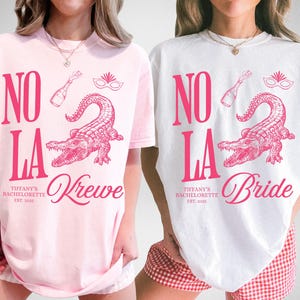 May include: Two t-shirts, one pink and one white, with a pink graphic design. The design includes the words "NO LA", an alligator, a champagne bottle, and a mask. The pink shirt says "Krewe" and the white shirt says "Bride".