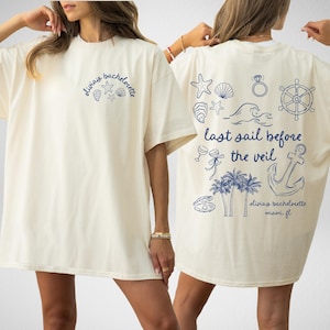 Peut inclure: Deux t-shirts blancs avec du texte et des graphismes bleus. Le devant d'un t-shirt dit "Olivia's Bachelorette" avec une étoile de mer, un coquillage et une ancre. Le dos de l'autre t-shirt dit "Last Sail Before The Veil" avec un coquillage, une ancre, des palmiers et le texte "Olivia's Bachelorette Miami, FL".