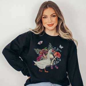 Puede incluir: Sudadera negra con un ganso blanco rodeado de setas, hojas y mariposas de colores.