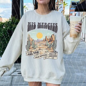 May include: Cream-colored sweatshirt with a desert landscape graphic. The design features cacti, a setting sun, and the text "HIS MERCIES ARE NEW EACH MORNING". The sweatshirt has a crew neck and long sleeves.
