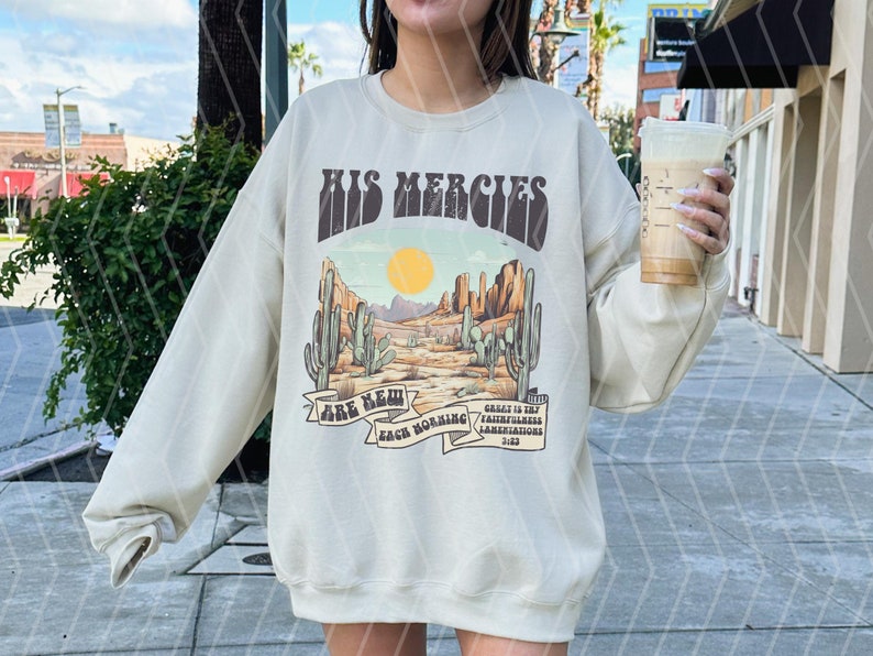 May include: Cream-colored sweatshirt with a desert landscape graphic. The design features cacti, a setting sun, and the text "HIS MERCIES ARE NEW EACH MORNING". The sweatshirt has a crew neck and long sleeves.